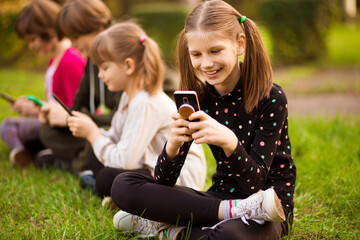 Fototapeta premium Full length photo of outdoor portrait of positive inspired child, girl sitting on grass with legs crossed and using mobile phone in company of friends