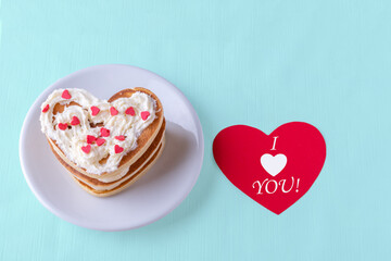 Valentine's day creative breakfast. Homemade heart shaped pancake decorated with white cream with red hearts on a white plate on a blue background with a red heart with the inscription I love you
