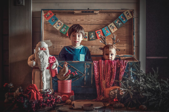 Cute little boy and his funny baby sister in deer antlers hat are playing in old vintage chest full of retro christmas stuff. Image with selective focus, noise effects and toning