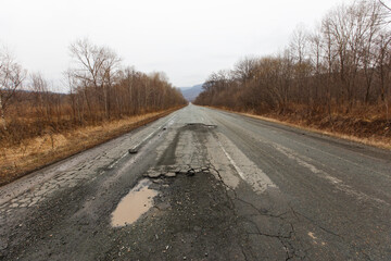 Bad Russian asphalt road. The asphalt road is full of holes and cracks among dried autumn trees.