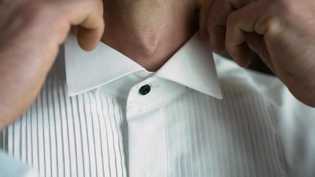 Caucasian young man stands indoors by the window and elegantly adjusts the collar of a white shirt with black buttons with clean neat hands with a watch