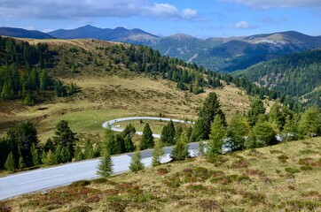 Naklejka premium Alpine road called Nockalmstrasse (Nockalm Road) in Kaernten (Carinthia), Austria, mountain range of Nockerberge (Nocky Mountains) on horizon, sunny autumn landscape, blue sky with clouds background 