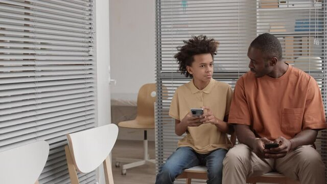Medium long of young African man and school boy dressed casually using their smartphones, talking, sitting in waiting room