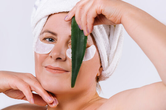 Portrait Of Caucasian Elderly Woman With Fair Skin, Cosmetic Eye Patches And Aloe Vera Leaf In Front Of Face, Closing An Eye. White Background.