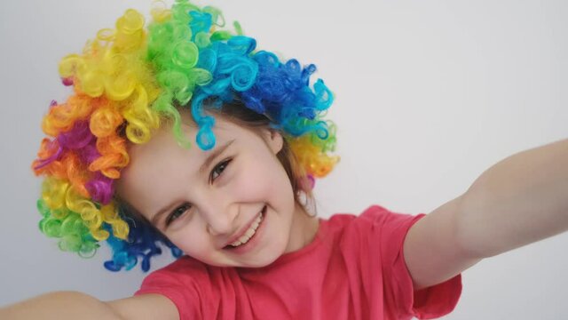 Portrait of cheerful little girl in rainbow wig on white wall background