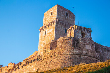 Fototapeta premium Close up view of medieval castle up the hill of Assisi, province in Umbria Italy. Stone bricks tower and walls at evening with clear clouds sky 