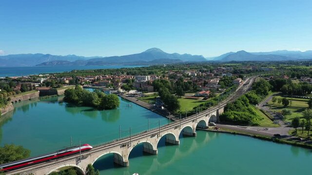 Frecciarossa high-speed train passes an arched railway bridge over a river in the background Lake Garda Italy. Red high-speed train on the bridge