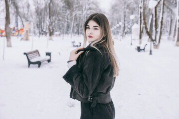 Fototapeta na wymiar A beautiful young girl in warm clothes walks, plays and poses against the background of a winter snow-covered park and a playground