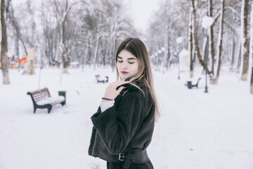 Fototapeta na wymiar A beautiful young girl in warm clothes walks, plays and poses against the background of a winter snow-covered park and a playground