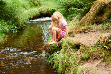 Child cute blond girl playing in the creek. Gril walking in forest stream and exploring nature. Summer children fun. Children summer activities - Powered by Adobe