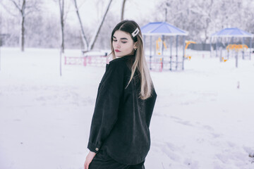 A beautiful young girl in warm clothes walks, plays and poses against the background of a winter snow-covered park and a playground