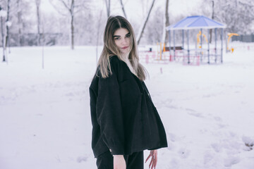 A beautiful young girl in warm clothes walks, plays and poses against the background of a winter snow-covered park and a playground