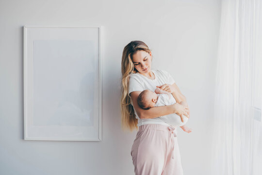 Careful mother with long loose flowing hair cradles baby holding daughter in arms and standing near large picture on wall under bright light at home.