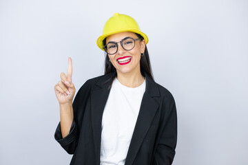 Young architect woman wearing hardhat showing and pointing up with fingers number one while smiling confident and happy