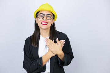 Young architect woman wearing hardhat smiling with her hands on her chest and grateful gesture on her face.