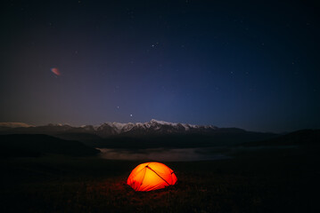 Atmospheric mountain landscape with vivid orange tent on hill under night starry sky. Tent glow by orange light with view of snowy mountains in starry night. Overnight in nature in great mountains.