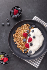 Homemade granola with yogurt and fresh berries in black ceramic plate on dark stone background. Vertical photo - Image