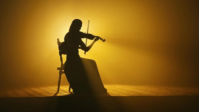 Silhouette of a Girl violinist in a Long Dress. A Musician girl Sits on the Stage and Performs a Classical Piece of Music on the Violin. Scene on a Dark Background in Smoke with Spotlights.