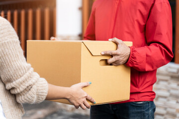 An Asian deliver man in red uniform handing parcel to a female costumer in front of the house. A postman and express delivery service deliver parcel during covid19 pandemic.