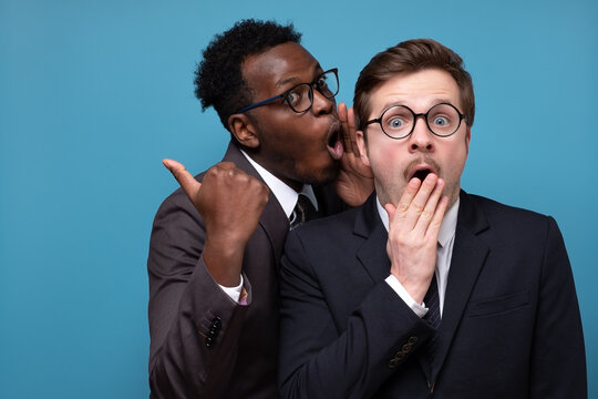 African man whisperingto his caucasian coworker ear telling him something secret. Studio shot on blue wall. Human emotions facial expressions