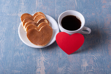Homemade heart shaped pancakes on a white plate, a mug with coffee or cocoa and a red blank heart shape on a blue stylish wooden background, top view. Creative breakfast for Father's Day. Love concept