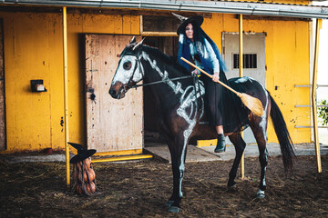 A girl dressed as a witch with a broom sits on a horse on which a skeleton is drawn