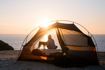 Woman and her pet dog sit at beach next to tent, solitude camping in nature. Sunset shot of young woman have self exploration time and connection to nature. Tranquility and mindfulness
