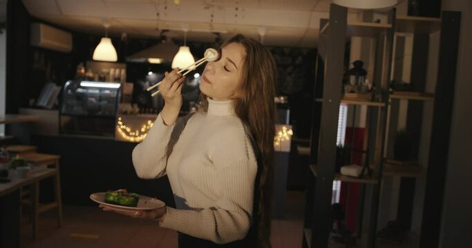 A Beautiful Young Long-haired Girl Sits On A Table In A Cafe With A Plate Of Sushi In Her Hands And Holds One Roll With Chopsticks At Eye Level