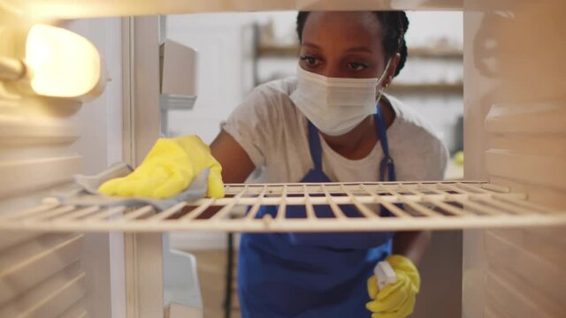 African maid in apron, gloves and safety mask cleaning refrigerator