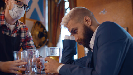 Bartender in safety mask cleaning counter full of empty glasses and tired businessman drinking beer