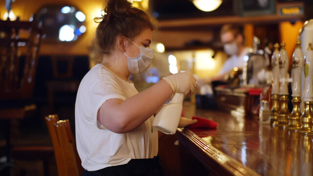 Young waitress in safety mask wiping bar counter with duster and detergent