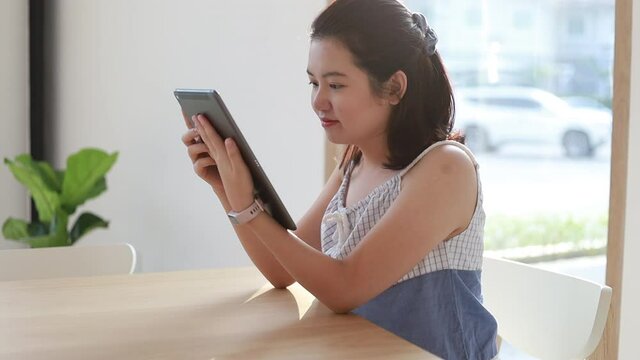 Young Asian woman is surfing the internet on a Tablet sitting in a coffee shop smiling, Have fun and enjoy, Use the internet at 5G speed, 4K Footage.