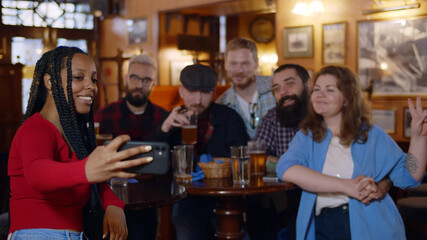 Charming excited african woman taking selfies with her friends at beer pub.
