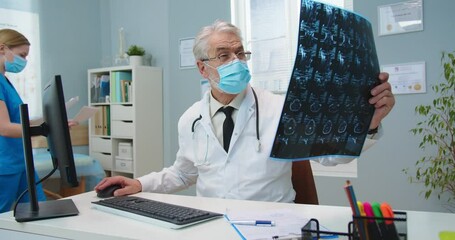 Smart Caucasian male doctor in medical mask looking at MRI scan in hospital office. Senior man in white gown comparing x-ray with computer reports. Medicine, pandemic, technology concept. - Powered by Adobe