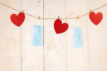 Red hearts and face masks hanging on cord on white wooden background. Backdrop with valentine tematics during pandemic of coronavirus. Love symbol and surgical masks on wooden desk.