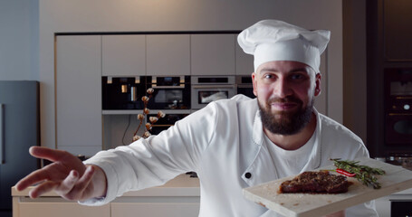 Young handsome chef in white uniform holding wooden board with ready beef steak