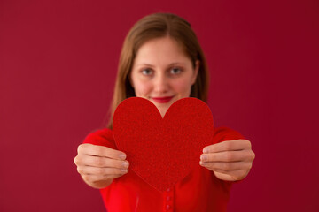 Blurred female showing heart to the camera on red background. Blonde girl holding glittered symbol of love on colorful backdrop. Smiling woman on valentine's day from close up.