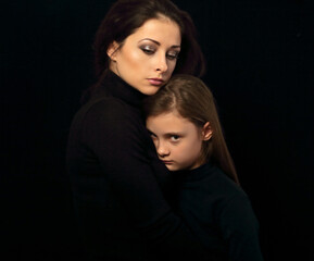 Serious thinking mother hugging her sad daughter on black background in dark shadow. Closeup family emotional portrait