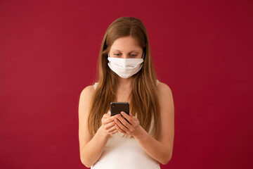 Woman in face mask texting on mobile phone with red background. Girl with respiratory protection sending messange on valentine's day. Concept of online dating during pandemic.