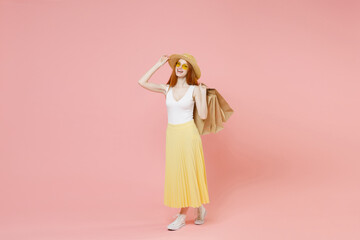 Full length body young happy excited redhead woman in straw hat glasses summer clothes look camera hold package bags with purchases after shopping isolated on pastel pink background studio portrait