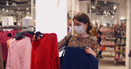 Fototapeta premium Young woman in medical mask choosing clothes in store