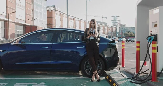 Woman stands with phone near her electric car and waits when vehicle will charged. Woman Connecting A Power Supply Cable Into An Electric Car For Charging An EV Battery. Charging An Electric Car