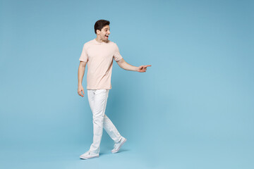 Naklejka premium Full length of young caucasian friendly smiling student man 20s in beige t-shirt white pants point index finger aside on copy space workspace area mock up isolated on blue background studio portrait.