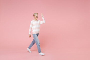 Fototapeta na wymiar Full length of smiling young friendly woman 20s short haircut in sweater jeans walk look aside waving and greeting with hand as notices someone isolated on pastel pink background studio portrait.