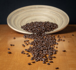 Simple yet refined backgrounded elegant composition of Italian dark roasted coffee beans falling out from a nude brown handmade wooden plate in the background towards foreground.