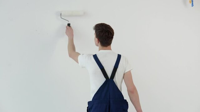Painter with a roller in his hands paints a white wall. View from the back