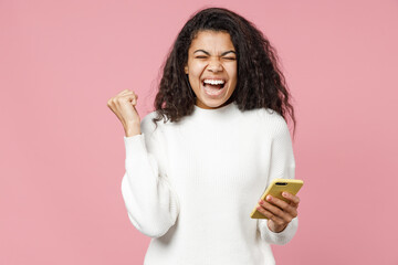 Young smiling attractive african american curly woman 20s wear white casual knitted sweater hold mobile cell phone do winner gesture clenching fist isolated on pastel pink background studio portrait.