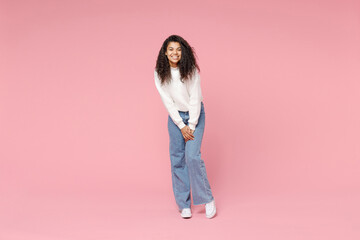 Full length young smiling cute african american woman 20s curly hair wear white casual knitted sweater jeans looking camera standing leaning over isolated on pastel pink background studio portrait. - Powered by Adobe
