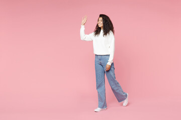 Full length young friendly smiling nice african american woman 20s curly hair wear white casual knitted sweater jeans walking waving hand greeting isolated on pastel pink background studio portrait.