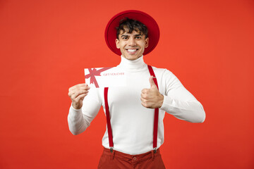 Young spanish latinos smiling man 20s in hat white shirt trousers with suspenders hold in hand gift voucher flyer mock up showing thumb up like gesture isolated on red color background studio portrait
